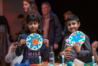 Two kids smiling and holding up colourful paper plates they have decorated.