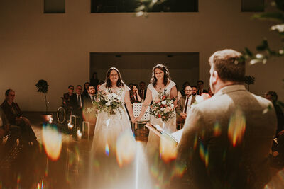 Two brides stood together holding hands in a candle lit room surrounded by friends and family.