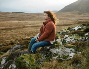 Woman with red hair wearing a rust coloured coat, blue jeans and green wellies sat in some grassy hills.