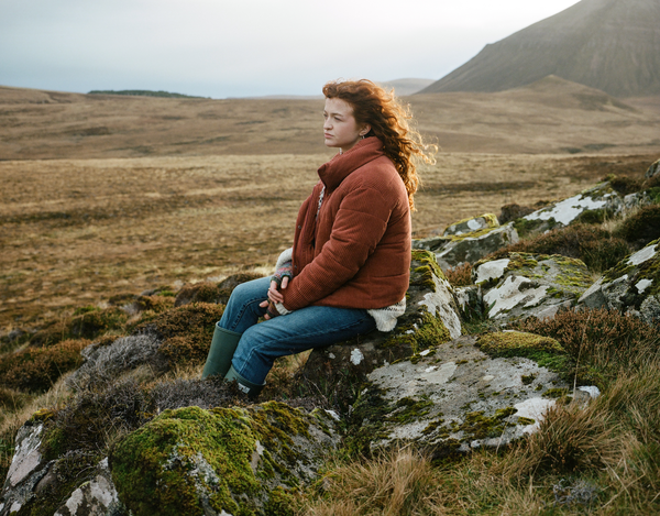 Woman wearing a rust coloured coat, blue jeans and green wellies, sat in some grassy hills.