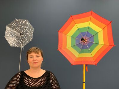 Artist with artwork, two sideway umbrellas on poles. One polka dot, the other rainbow. 