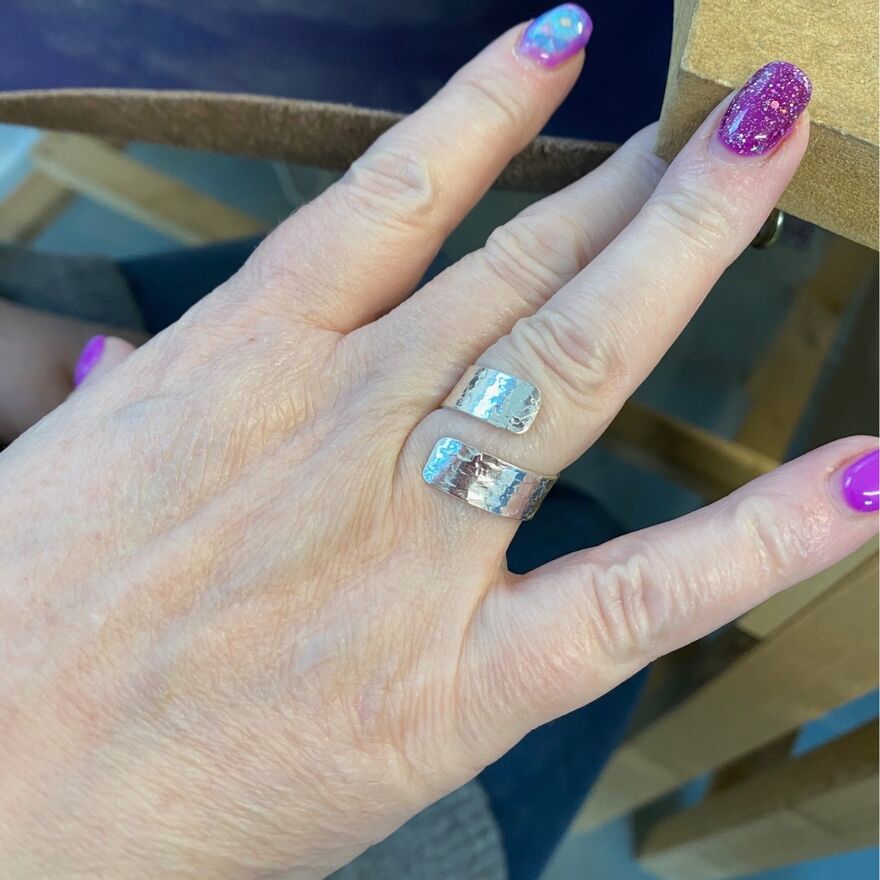 A ring made from a flat unconnected piece of silver sheet that overlaps is wrapped around the finger of someone with sparkly purple nails.