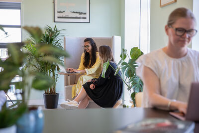 Two women sat together looking at a laptop and smiling, In the foreground there are plants and another woman working at a laptop, wearing glasses and a white top.