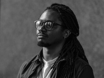Artist portrait of Larry Achiampong. Larry is Black with dreadlocked hair, a short beard, glasses and a dark shirt.