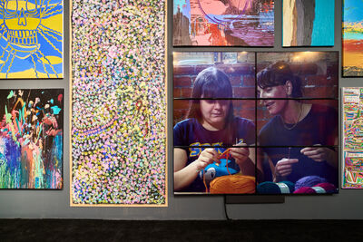 Dark grey wall with big, colourful canvases on it showing abstract art. There is also a large video screen showing two women sat side-by-side, knitting bright coloured with wool.