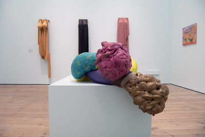 A white room with a sculpture in the middle of a colourful fabric tube tied in a knot with expanding foam leaking of the ends. There are three silk dresses displayed on the wall in the background.