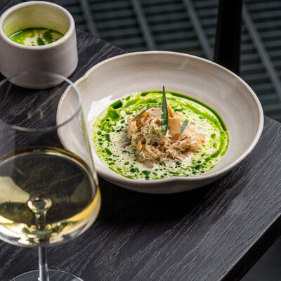 White bowl with fish surrounded by a bright green sauce in it. A glass of white wine is next to the dish.