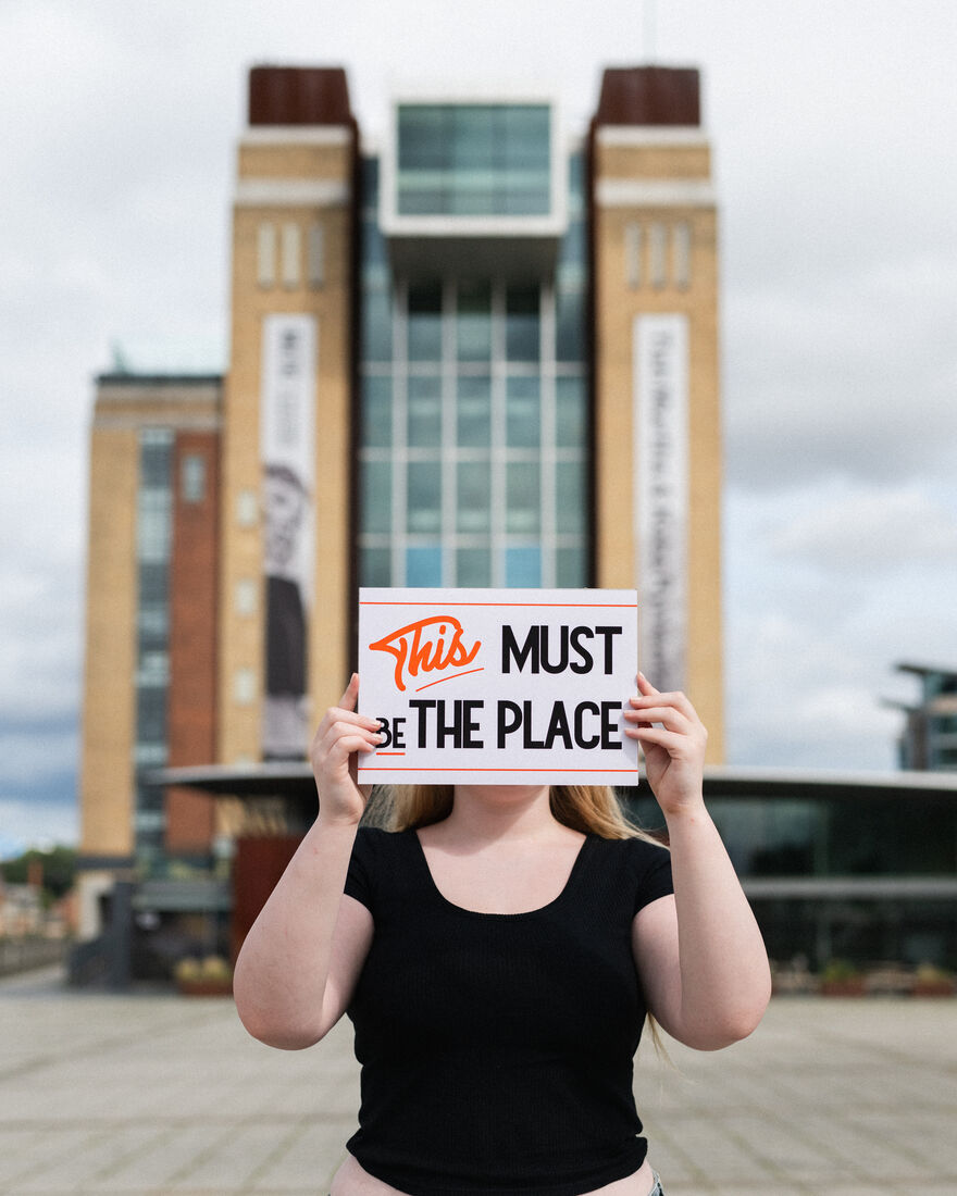 Person holding up a landscape print that reads 'This Must Be The Place'