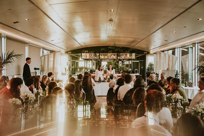 A full restaurant with two brides at the top table giving a speech and smiling.