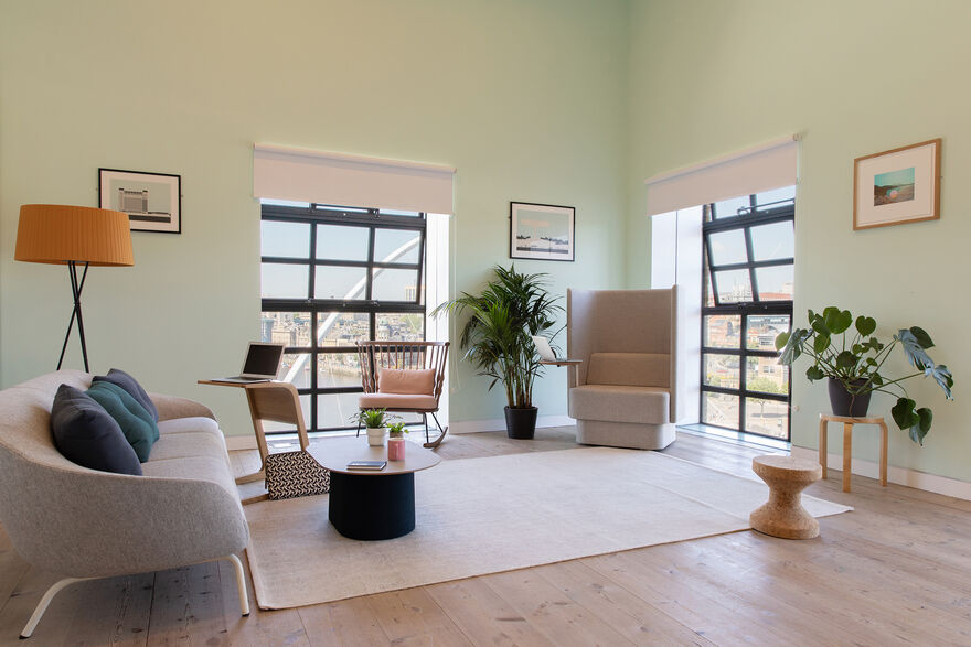 A light green room with big windows and a large shared desk with seats. There are green plants in the background and foreground.