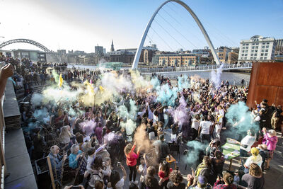 Crowd of people with hands in the air throwing brightly coloured cloud paint. Behind them is the River Tyne blue on a sunny day.
