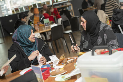 Two women wearing headscarves, sat at a table, crafting and talking.