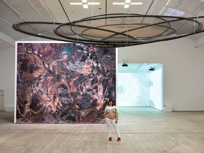Artist stood in exhibition with large artwork behind and circular nets hanging above