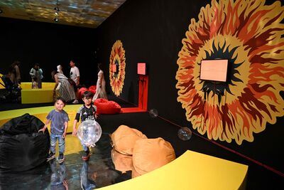 A colourful and abstract room with yellow, orange and black colours and bean bags on the floor. There are two small children running towards the camera chasing a floating glittery bubble.