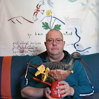 Man wearing a blue top and sat on a blue sofa holding a red pot with a model of a bird sat on it.