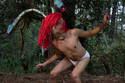 Man wearing just white pants, a mask and red felt hair stood in a forest.
