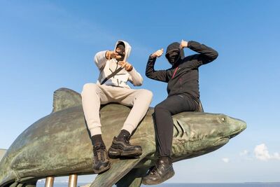 Two young people in black and white tracksuits wearing ski masks. They sit on a shark sculpture at the sea side and hold their thumbs up and point at the camera.
