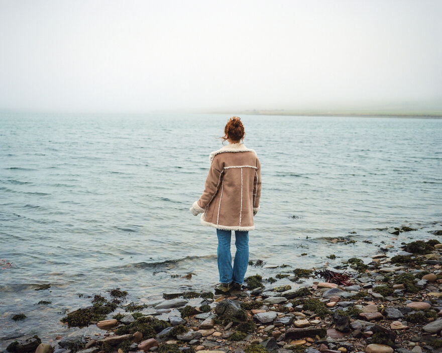 Woman with curly red hair wearing a big brown coat and looking out to sea.
