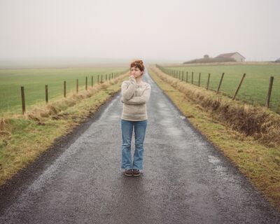 Woman wearing a grey jumper and blue jeans stood in the middle of a country road.
