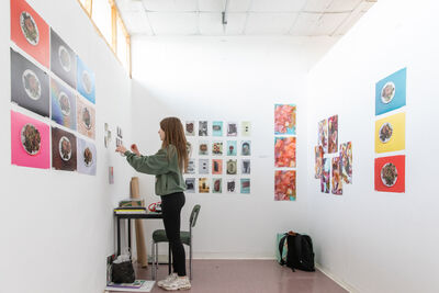 Woman wearing black leggings and a green hoodie stood in a small white room placing colourful artwork on the walls.