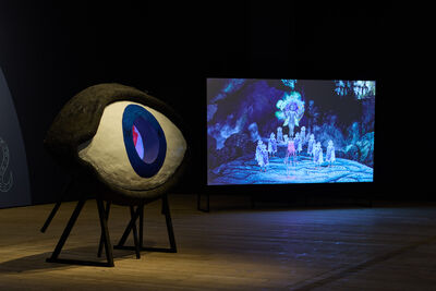 A screen showing an animation with a large sculpture of an eye next to it.