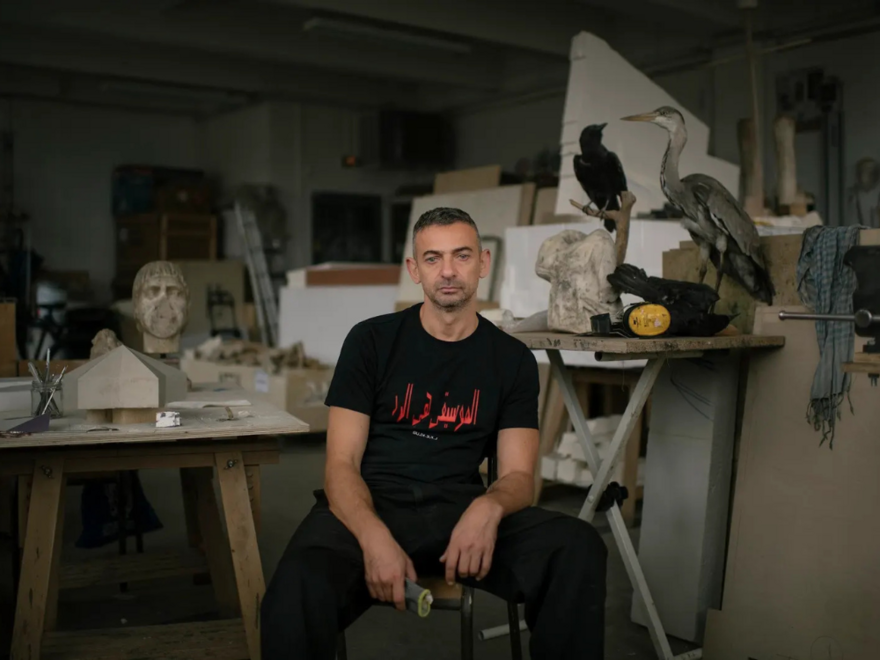 A man wearing a black t-shirt and black trousers sat on a chair in the middle of an art studio surrounded by sculptures.