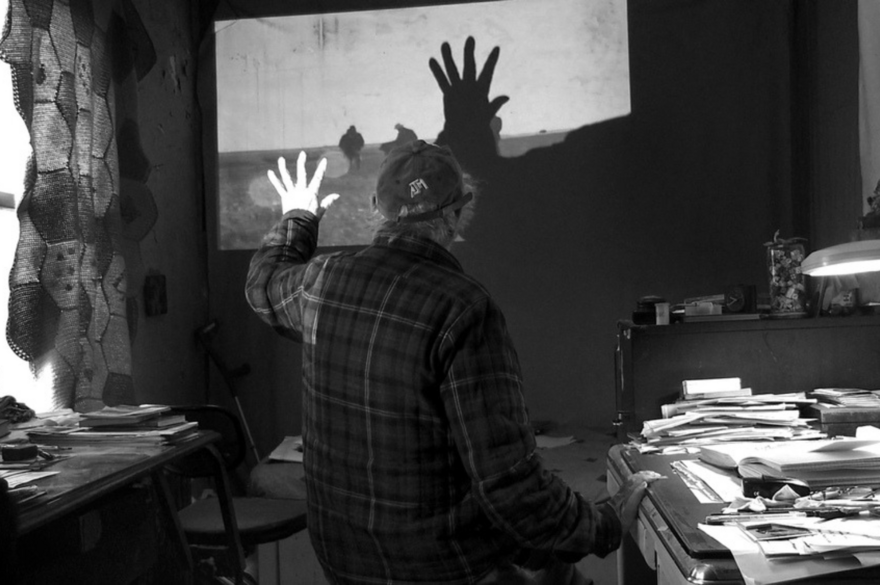 A person with their back to the camera holds their hand up wi a projected image on a wall in a room with paper and books scattered on a desk. The image is in black and white. 