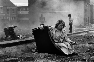 Black and white photo of a young woman sat on a sofa in the street, surrounded by smoke and rubbish.
