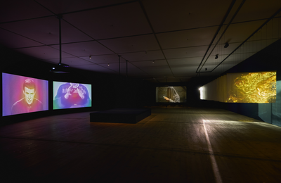 Dark room with large screens suspended from the ceiling and a big cushion seat in the middle of the room.