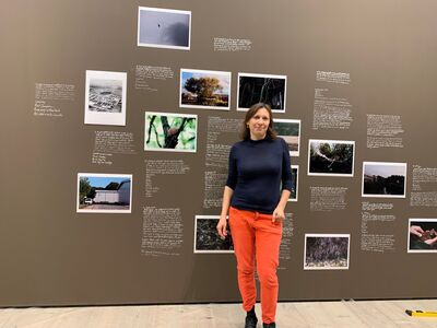 Artist wearing navy top and orange trousers in front of brown wall with photographs and handwritten text