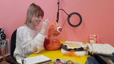 Young woman working on a felt sculpture, smiling with a pink wall in the background.
