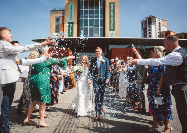 Two people in wedding attire smiling outside Baltic with a crowd of people throwing confetti. The sky behind is blue.