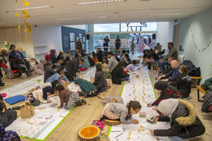 Room full of people kneeling and sat down drawing on long sheets of paper on the floor.