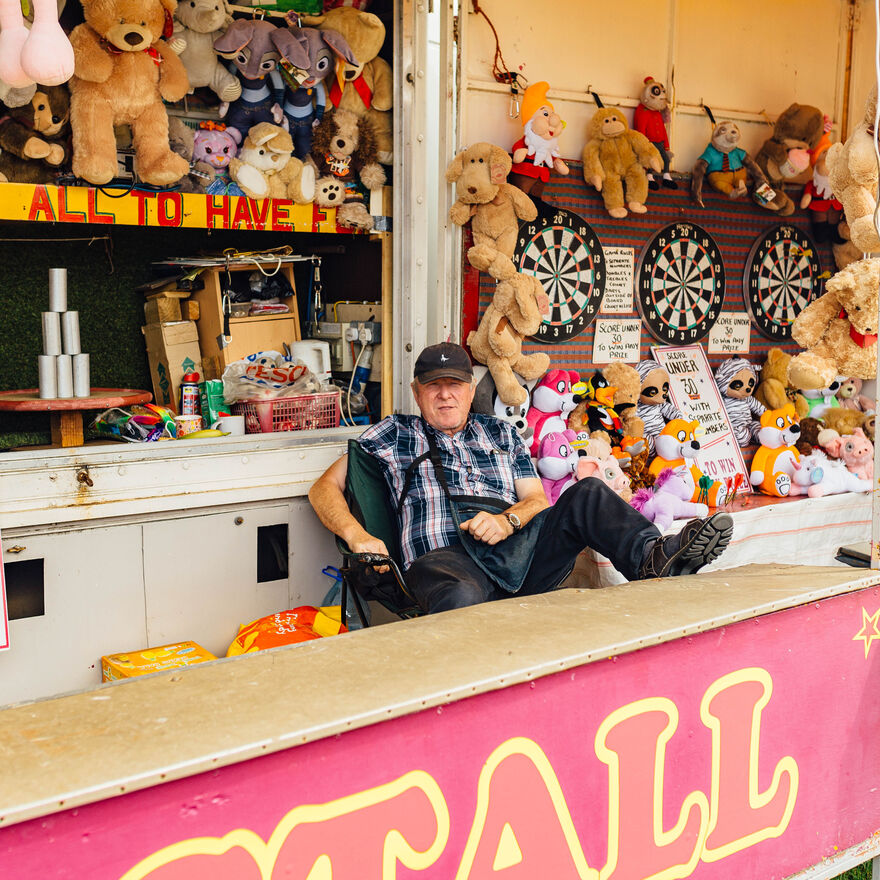 A man sits behind a stall with teddy bears and dart boards