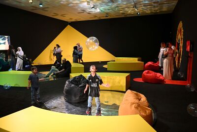 A colourful and abstract room with yellow, orange and black colours and bean bags on the floor. There is a small child in the middle of the room staring on wonder at a floating glittery bubble.
