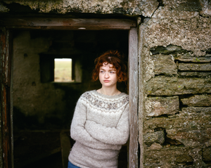 Woman with curly red hair and a grey wool jumper stood in a doorway to a barn.